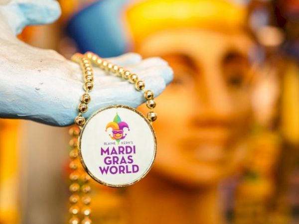 A hand holds a beaded necklace with a "Mardi Gras World" medallion, featuring a jester hat logo, against a colorful background.