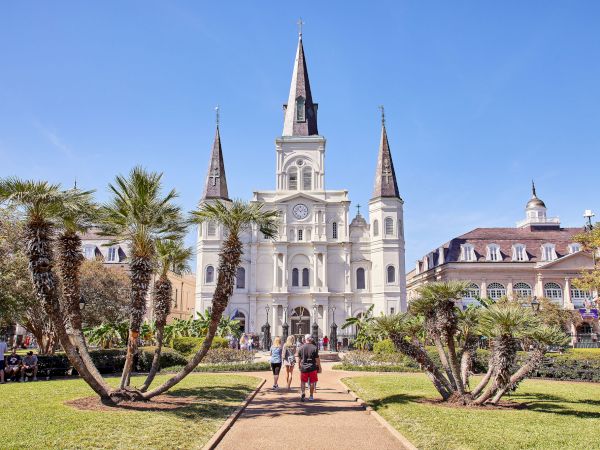 The image shows a cathedral with three spires, surrounded by palm trees and people walking on a pathway in a sunny park.