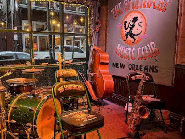 A jazz club stage with drums, a double bass, and a saxophone; banner reads "The Spotted Cat Music Club, New Orleans."