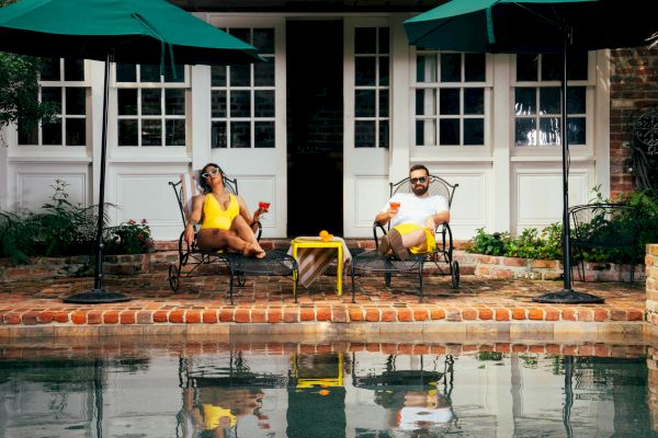 Two people relax on patio chairs by a pool, holding drinks. Green umbrellas provide shade, and a small table is between them.