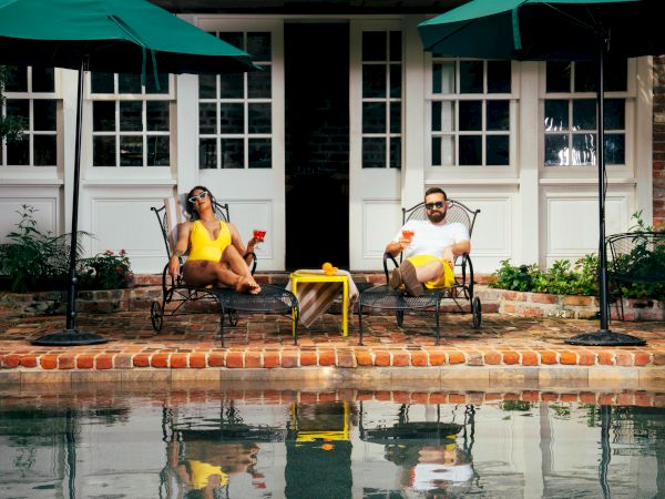 Two people relax on patio chairs by a pool, holding drinks. Green umbrellas provide shade, and a small table is between them.