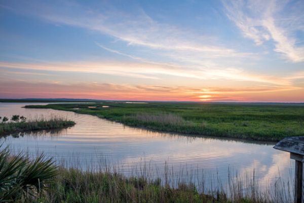 Sunset over a marshland with a winding waterway, grassy banks, and a colorful sky reflecting on the water.