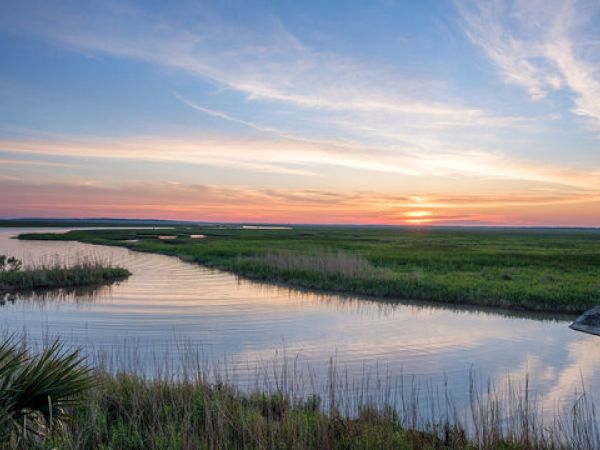 Sunset over a marshland with a winding waterway, grassy banks, and a colorful sky reflecting on the water.