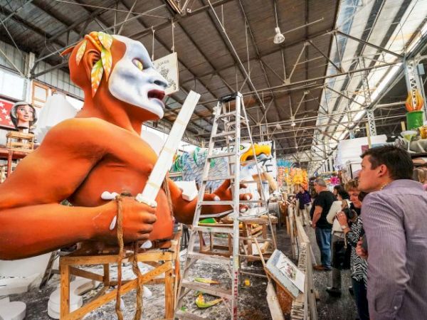 A man in a large orange mask sculpture is being painted in a workshop; sculptors work on a tall figure with a white head and sword, spectators watch, and ladders lean around.