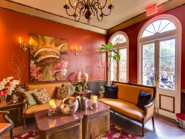 A cozy, elegant lounge with warm red walls, a chandelier, plants, and a leather sofa set around a wooden coffee table, near arched doors.