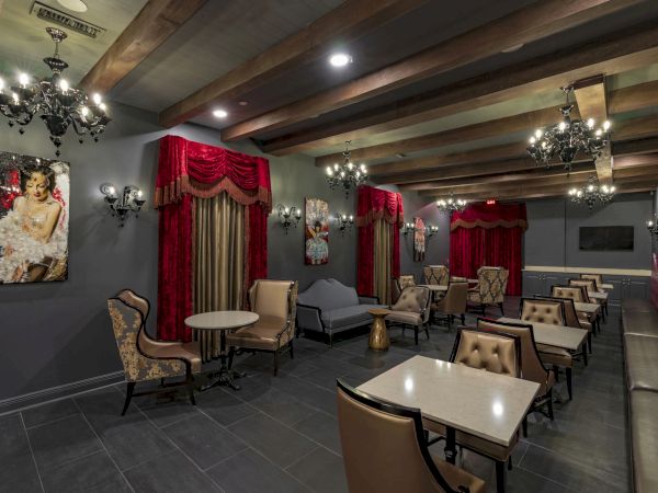 A stylish, dimly lit lounge with red curtains, chandeliers, framed art, and multiple tables and chairs arranged for upscale socializing.