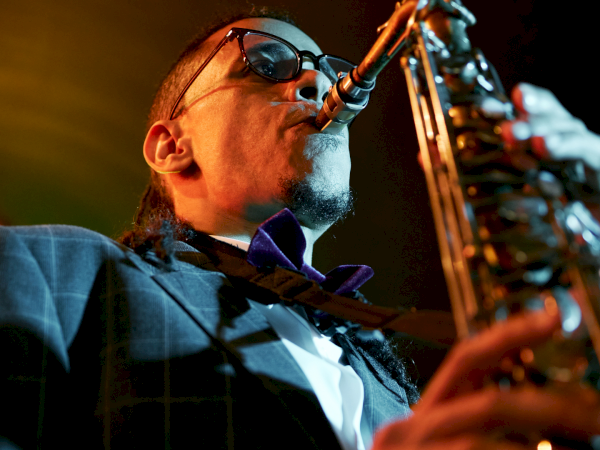 A stylish saxophonist in a suit and bow tie plays a sax solo under warm stage lights, wearing sunglasses as he performs.