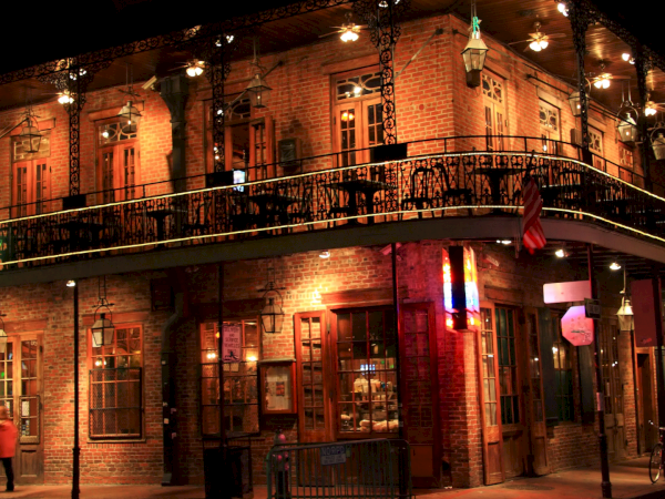 Old brick building with a second-floor balcony, warm lights, and a corner street scene at night. (140 chars)