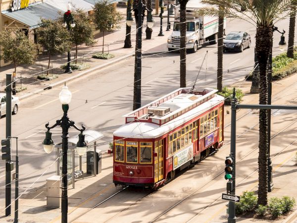 A vintage streetcar tram runs along city tracks beside palm trees, storefronts, and parked vehicles on a sunny street.