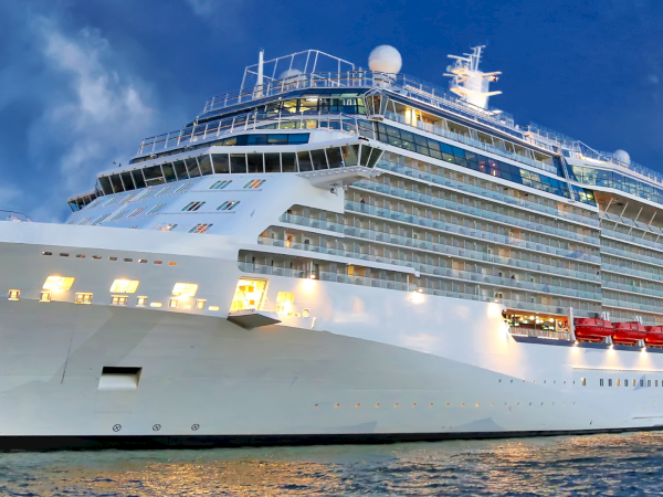 A massive white cruise ship docked at sunset, with multiple decks, balconies, and bright lights reflecting on calm water, ready for passengers.