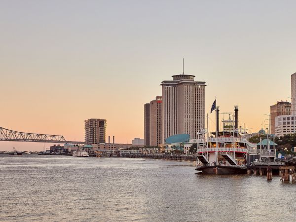 Riverfront skyline with tall towers and a docked paddlewheel boat along the water at sunset.