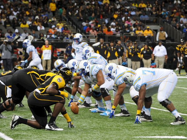 A football game with players from two teams crouched in a crowded pile during a snap, while the crowd watches in the stands.