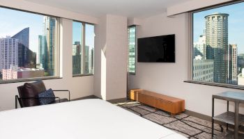 A modern hotel room with a bed, armchair, and TV. Large windows showcase a cityscape with tall buildings and a clear sky.