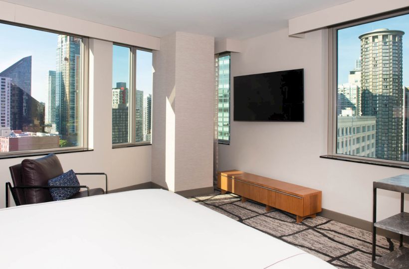 A modern hotel room with a bed, armchair, and TV. Large windows showcase a cityscape with tall buildings and a clear sky.