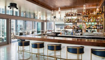 The image shows a modern bar with blue stools, a large selection of bottles, and floor-to-ceiling windows offering a city view.