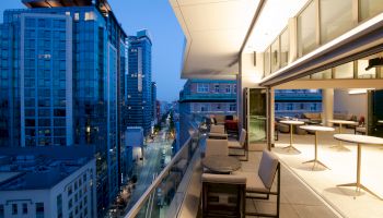 A modern balcony overlooking a city street with high-rise buildings, featuring outdoor seating and elegant lighting at dusk.