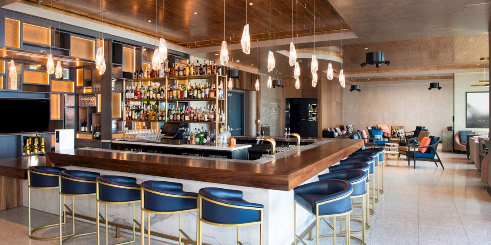 The image shows a modern bar with hanging lights, a stocked shelf, and stylish seating. There are tables and sofas in the background.