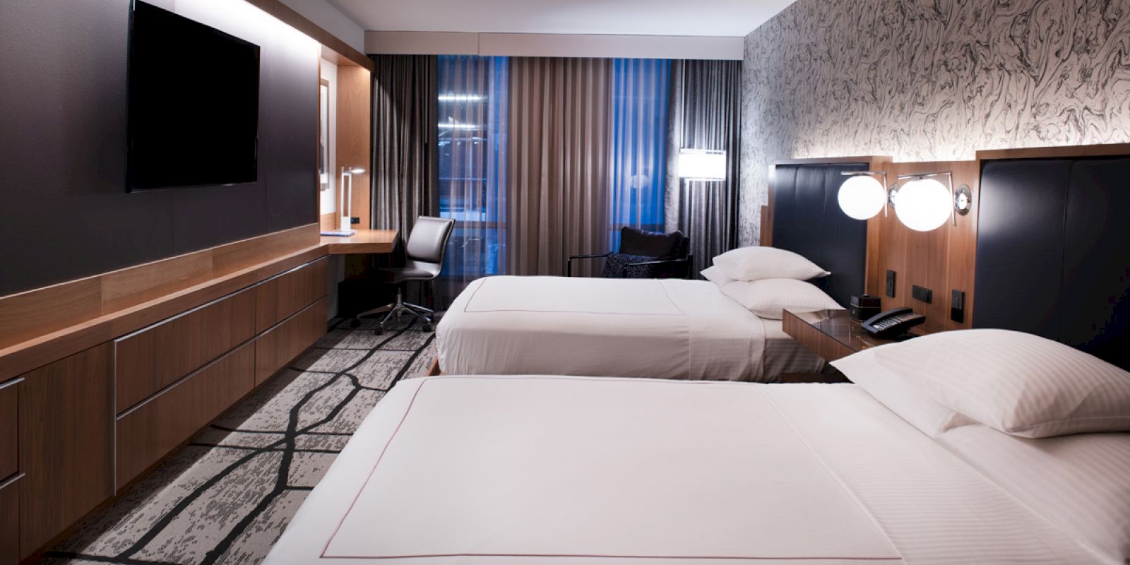 The image shows a hotel room with two beds, modern decor, wall-mounted TV, desk, chairs, and lamps on a patterned carpeted floor.