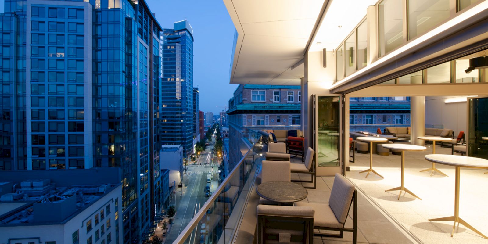 A modern balcony with outdoor seating overlooks a cityscape at dusk, featuring tall glass buildings and a street with lights.