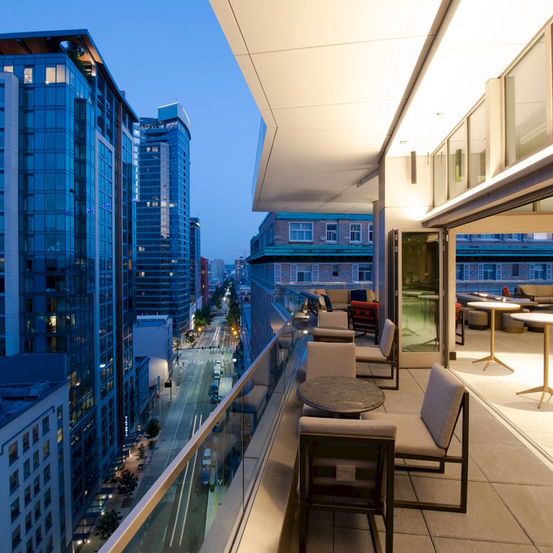 A modern rooftop terrace with tables and seating offers a view of a cityscape featuring tall, illuminated buildings at twilight.