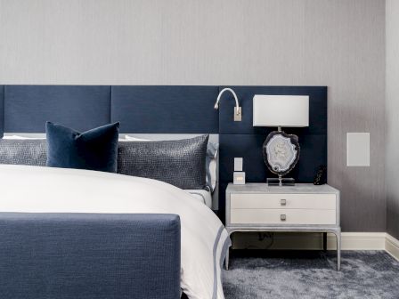 A modern bedroom features a blue headboard, white bedsheets, a bedside lamp, and a nightstand with drawers, creating a cozy atmosphere.