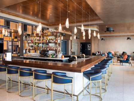 A stylish bar with a U-shaped counter, modern lighting, and shelves stocked with bottles; seating includes blue barstools.