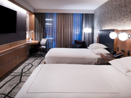 A modern hotel room with two beds, a wall-mounted TV, a work desk, and contemporary decor featuring a patterned accent wall.