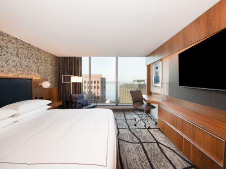 A modern hotel room with a king-sized bed, TV, desk, and a window view of city buildings. The decor is sleek and contemporary.