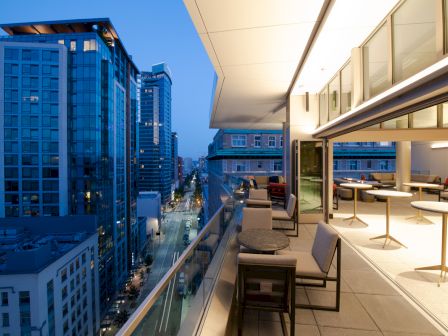 A modern cityscape at dusk shows tall buildings and a stylish balcony with tables and chairs overlooking the street below.