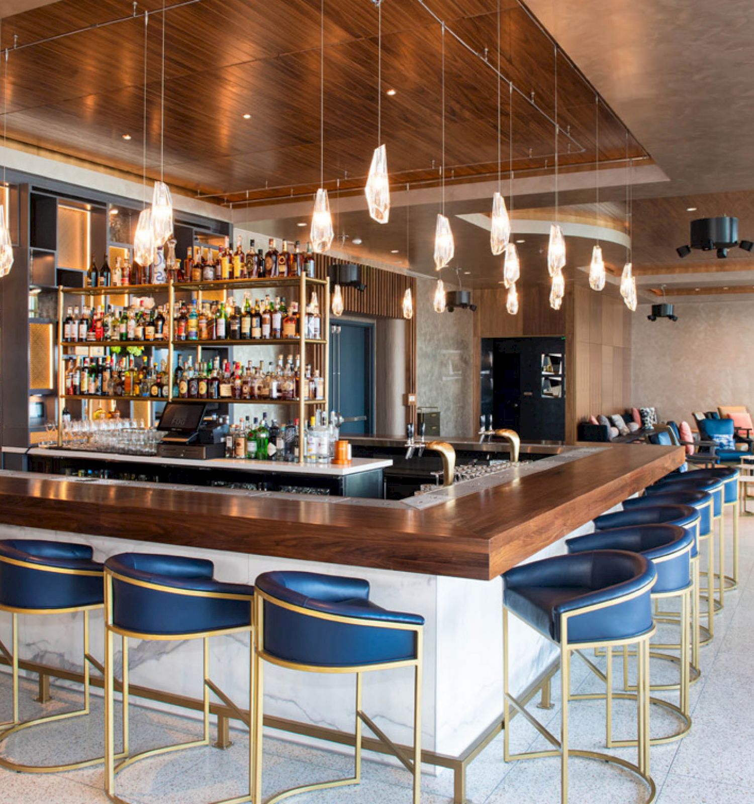 A modern bar with stylish blue stools, hanging lights, and shelves stocked with bottles. The space is open and inviting, with seating areas.