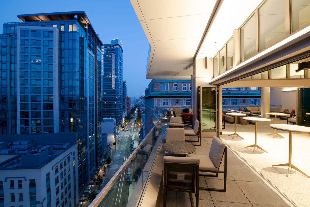 City view from a modern balcony with outdoor seating and buildings illuminated at dusk.