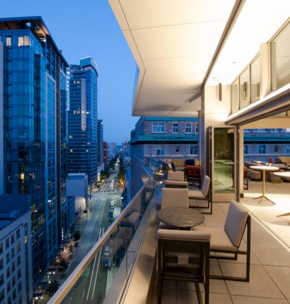 A modern balcony with tables and chairs overlooks a city street lined with tall, glass buildings at dusk.