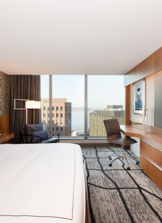A modern hotel room with a large bed, TV, desk, chair, and city view through floor-to-ceiling windows ending the scene.