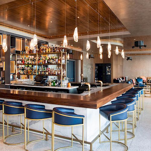 Sleek bar with a central counter, blue chairs, hanging lights, and a fully stocked display. Cozy seating area in the background.