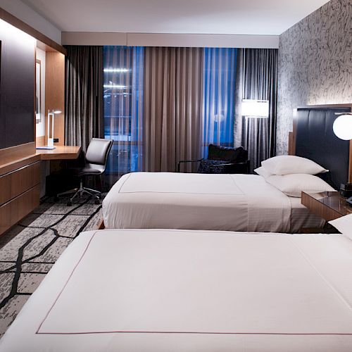 The image shows a modern hotel room with two beds, a TV, a desk, and a window with curtains, featuring stylish decor and lighting.