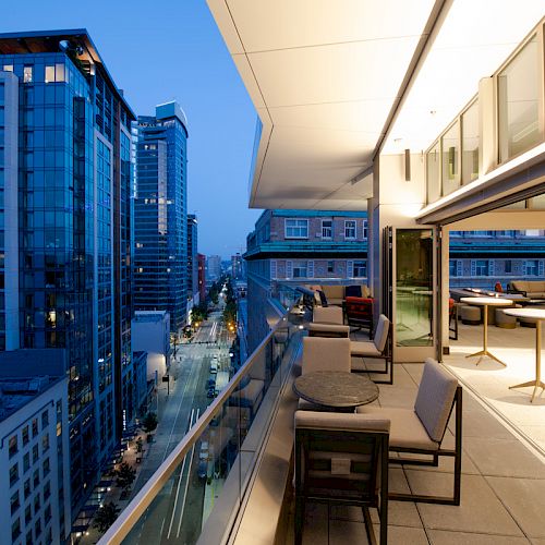A modern urban balcony overlooks a bustling city street at twilight, with seating areas and bright indoor lighting.
