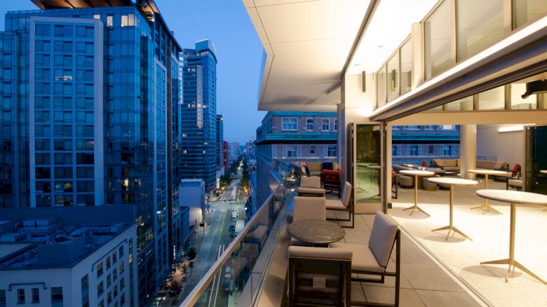 Rooftop terrace at dusk with tables and chairs, overlooking a cityscape of modern buildings and a street below.