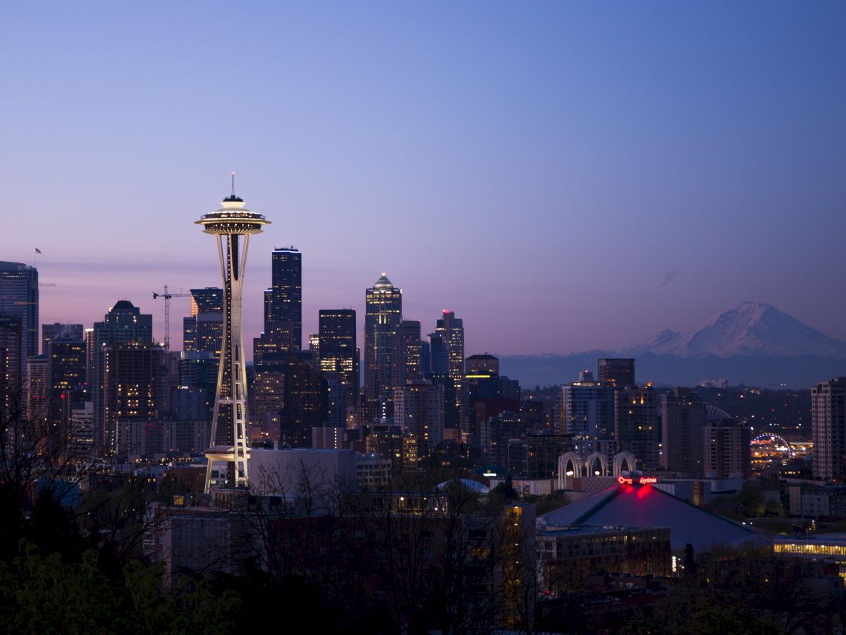 A twilight skyline of Seattle showing the Space Needle, city buildings, and Mount Rainier in the background under a clear blue-pink sky.