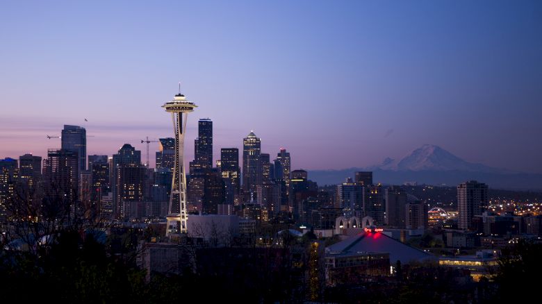 A twilight skyline of Seattle showing the Space Needle, city buildings, and Mount Rainier in the background under a clear blue-pink sky.