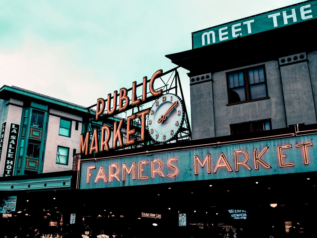 The image shows the entrance to a public market with neon signs reading “PUBLIC MARKET” and “FARMERS MARKET,” featuring a large clock.