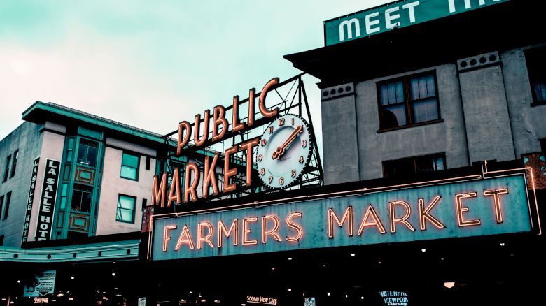 The image shows the entrance to a public market with neon signs reading “PUBLIC MARKET” and “FARMERS MARKET,” featuring a large clock.