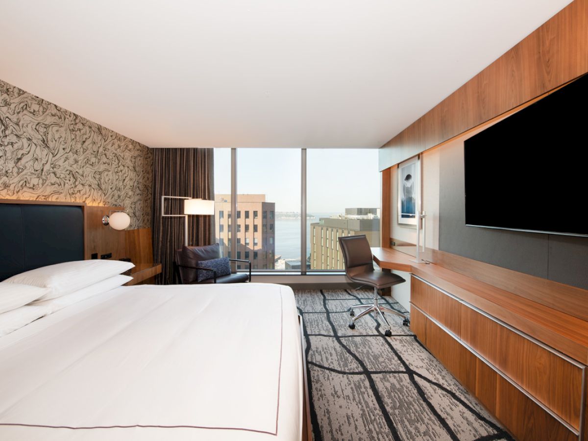 A modern hotel room with a bed, wall-mounted TV, desk, and a view of buildings through large windows.