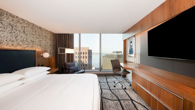 A modern hotel room with a bed, wall-mounted TV, desk, and a view of buildings through large windows.