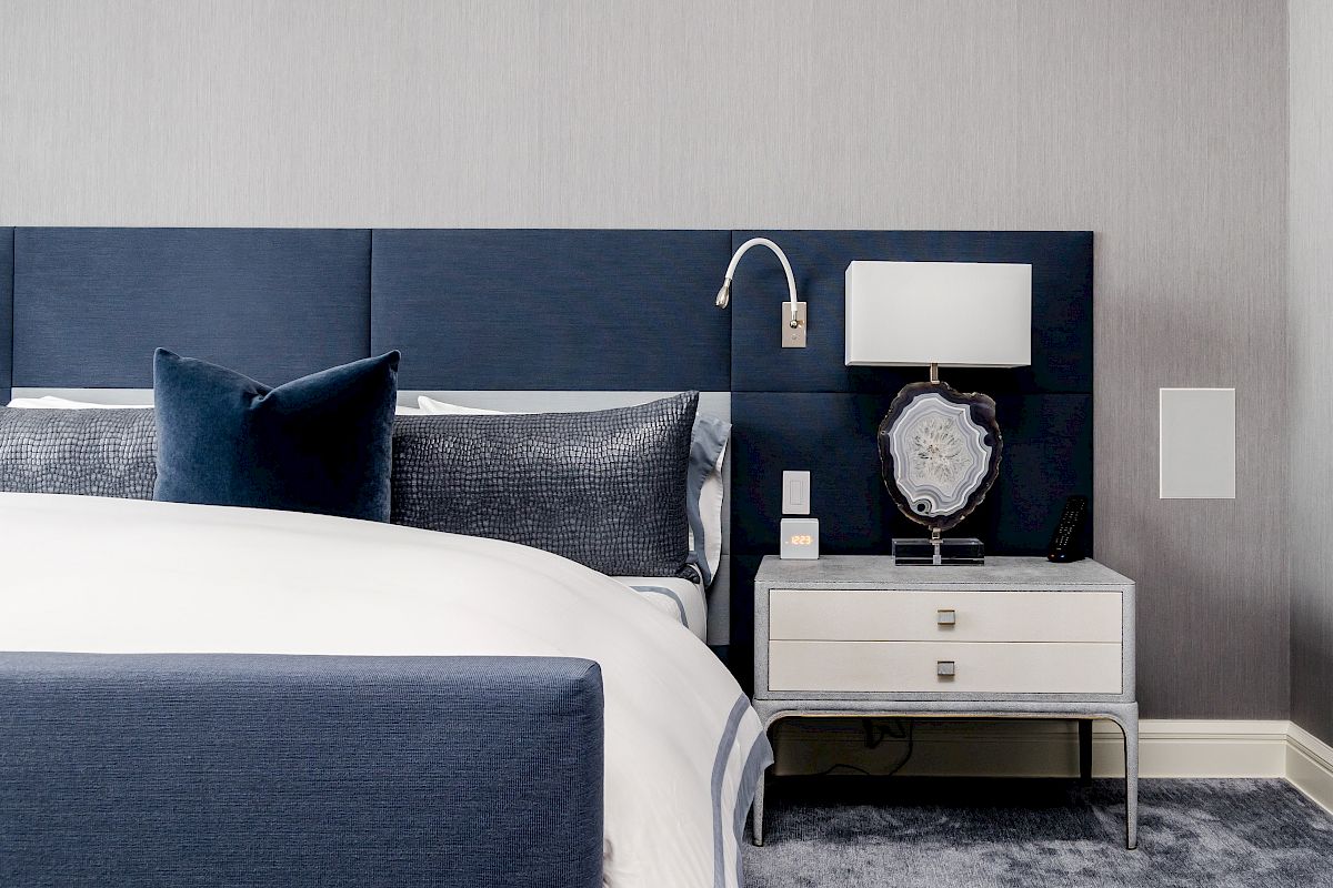 The image shows a modern bedroom with a blue upholstered bed, white linens, a bedside table with a lamp, and grey walls.