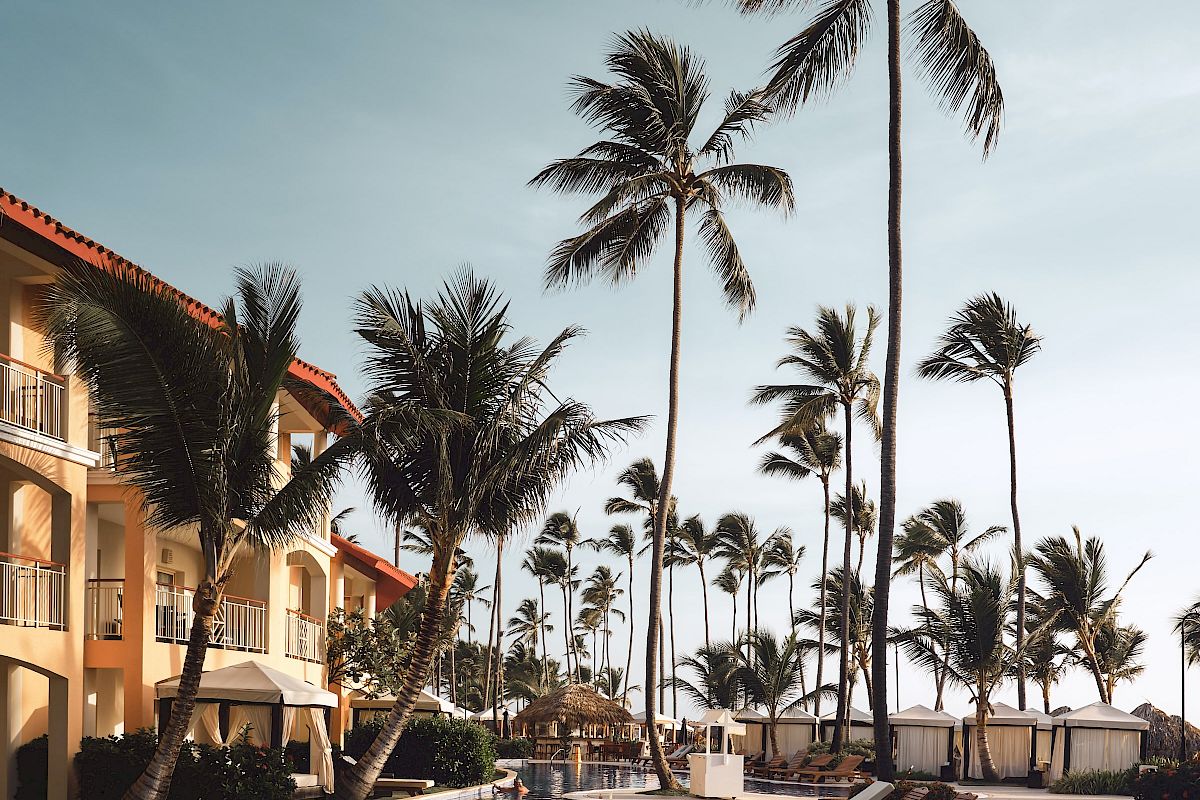 A serene poolside scene with lounge chairs, tall palm trees, and a warm building under a clear blue sky.