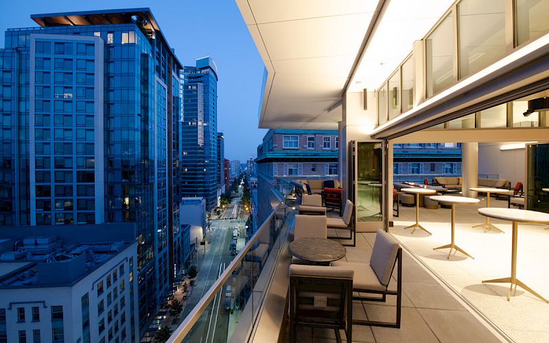A modern cityscape at dusk with high-rise buildings and an elegant balcony featuring tables and chairs overlooking a busy street.
