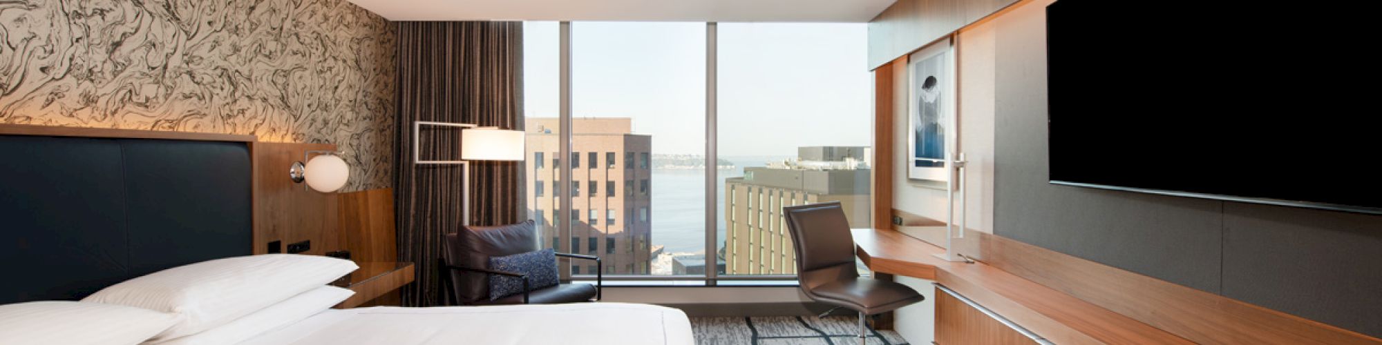 A modern hotel room with a bed, large TV, desk, chair, and window view of cityscape; décor features wood finishes and patterned walls.